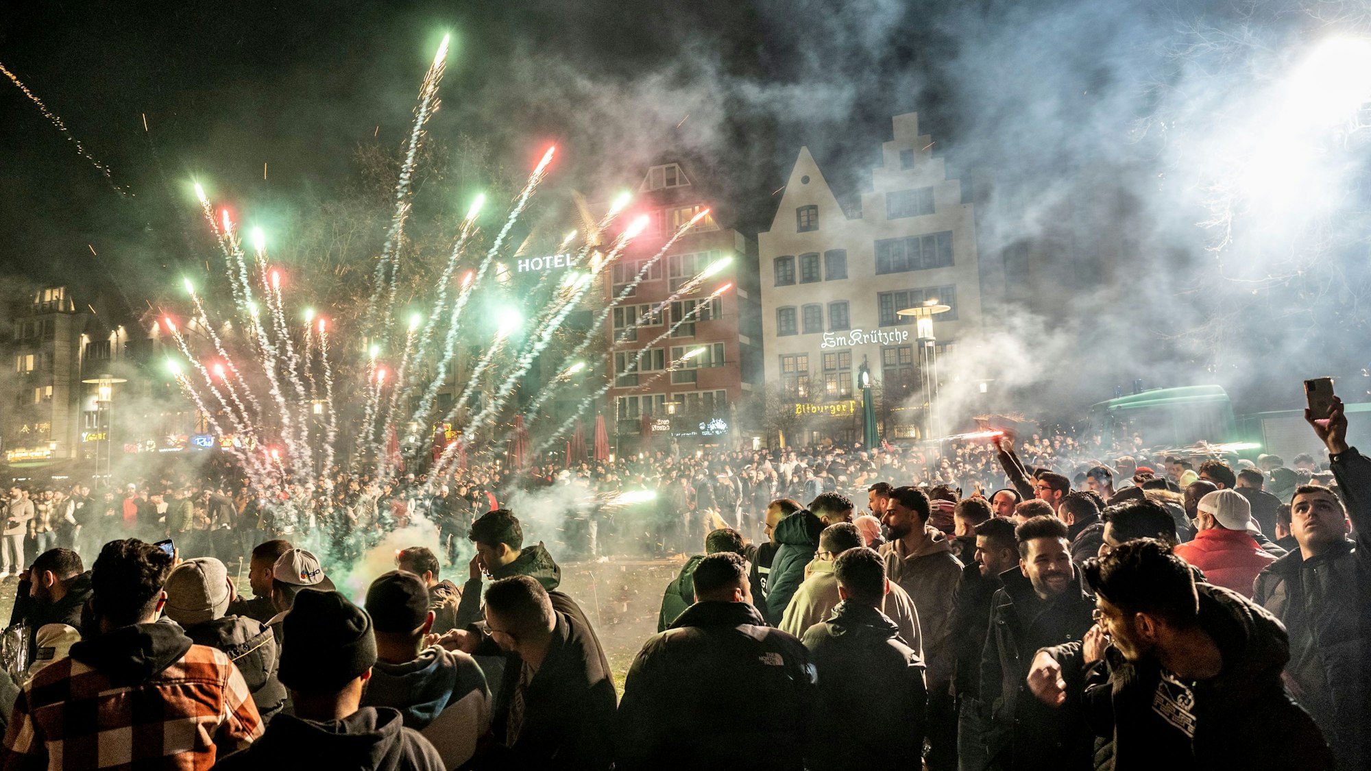 Silvester 2022 in der Kölner Innenstadt: Menschenmassen feiern in der Altstadt, in der Mitte sprüht eine Feuerwerksbatterie Funken und Rauch.