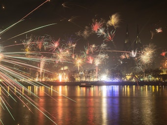 Feuerwerk steigt in den Nachthimmel und spiegelt sich im Wasser des Rheins.