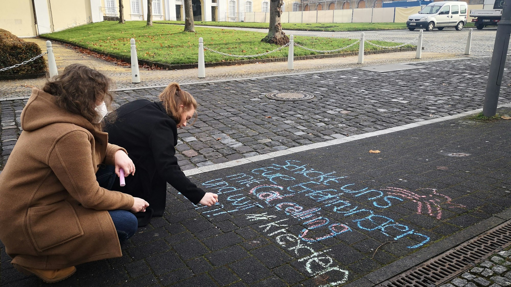 Zwei junge Frauen schreiben mit bunter Kreide einen Aufruf auf den Fußweg, mit dem sie dazu auffordern, Erfahrungen von Belästigungen in der Öffentlichkeit zu teilen.