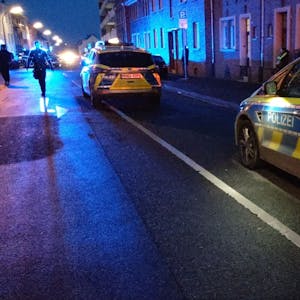 Zwei Streifenwagen stehen hintereinander auf einer Straße, man sieht mehrere Polizisten im Hintergrund. Die Straße ist dunkel, bis auf Laternen und Blaulicht.