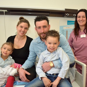Zu sehen ist eine fünfköpfige Familie mit einem Neugeborenen und der Hebamme in einem Krankenhauszimmer.