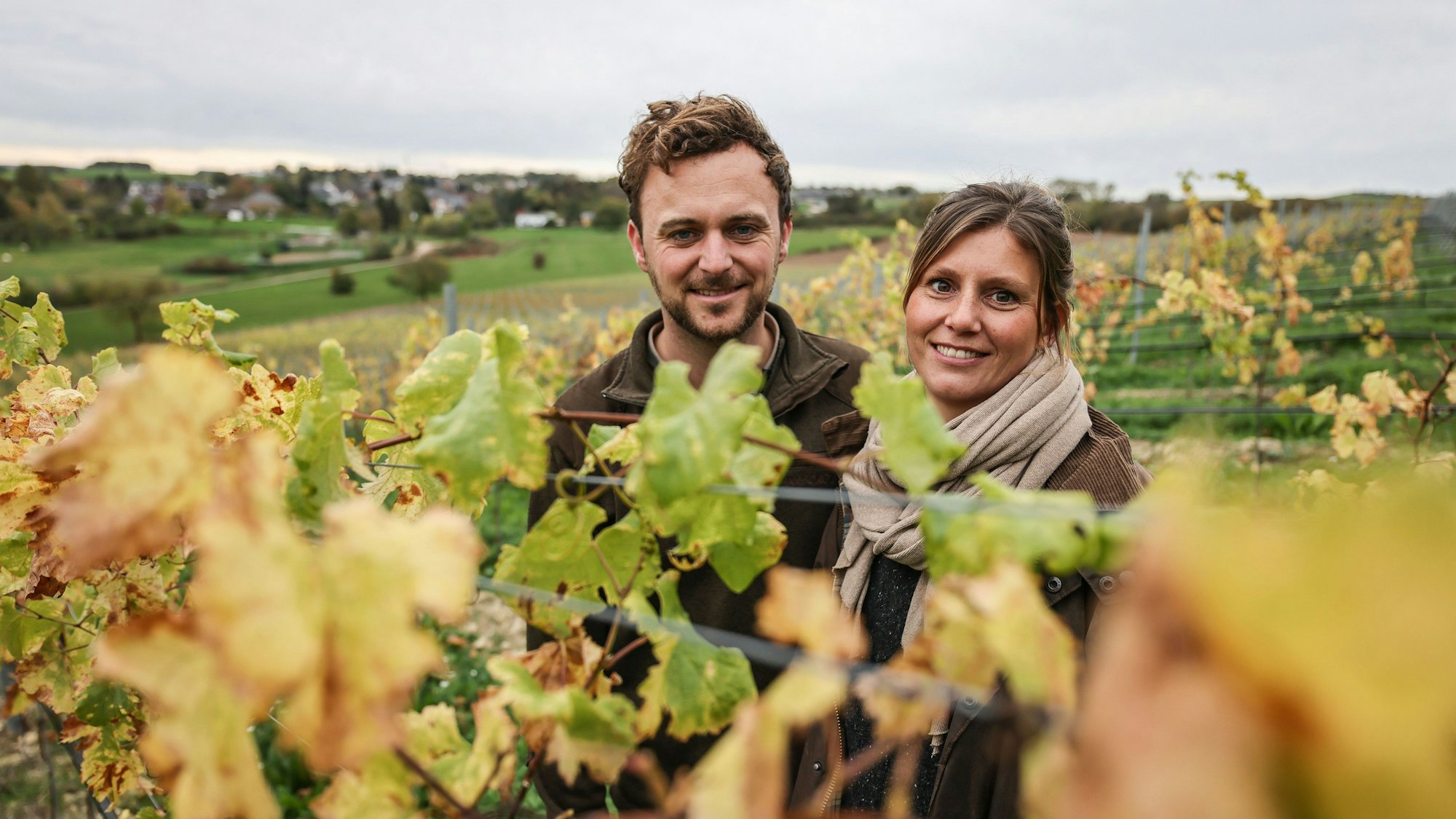 Ein junges Ehepaar steht inmitten von grünen und grün-gelben Weinreben in einem Weinberg.