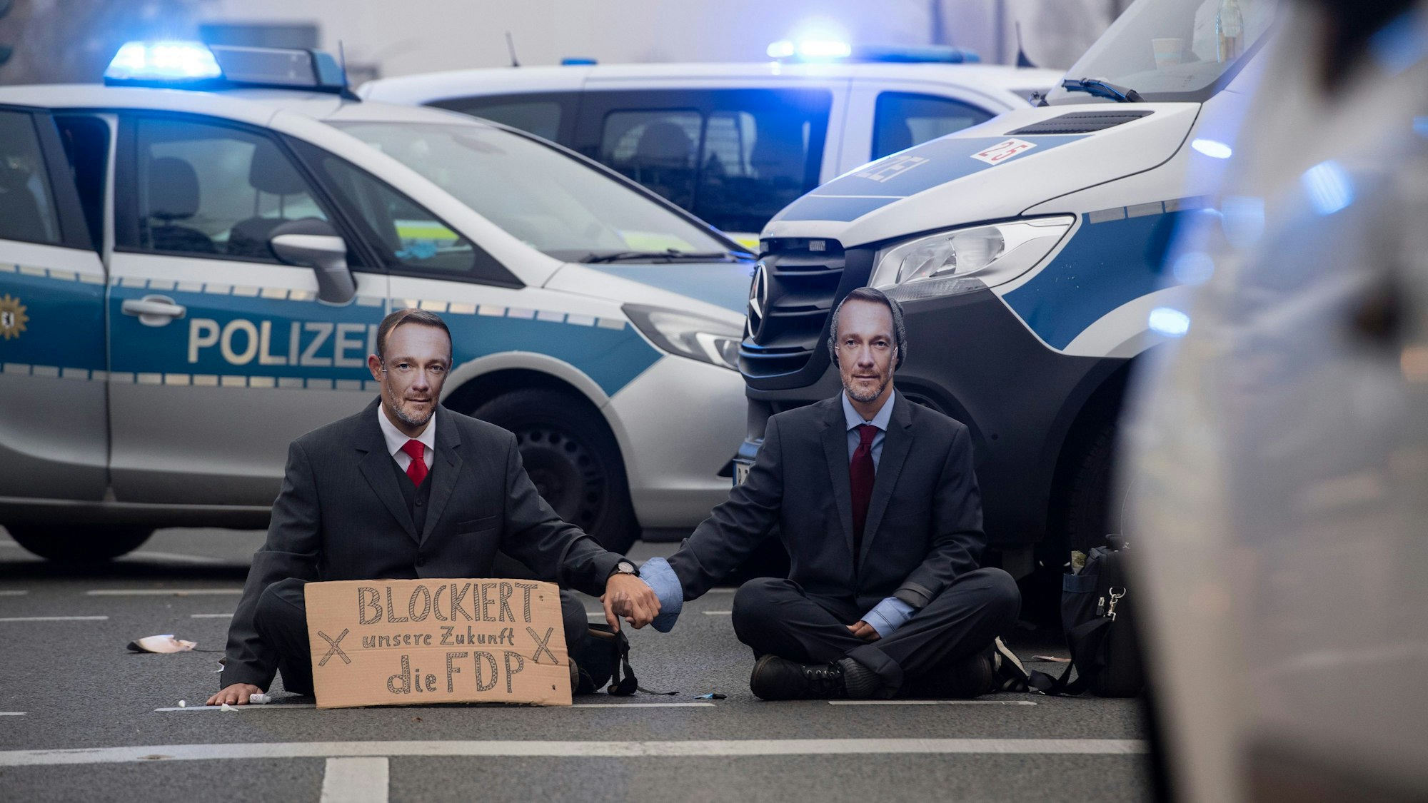 Aktivisten der Gruppe „Letzte Generation“ sitzen auf einer Kreuzung an der Landsberger Allee und tragen Masken, die Bundesfinanzminister Lindner zeigen.