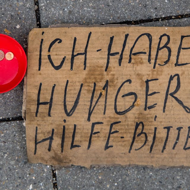 Ein dreckiges Pappschild mit der Aufschrift Ich habe hunger Hilf - bitte, liegt auf der Straße. Daneben steht eine rote kleine Schüssel mit zwei Münzen drinnen.