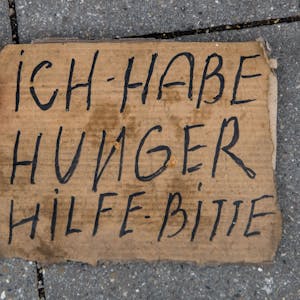 Ein dreckiges Pappschild mit der Aufschrift Ich habe hunger Hilf - bitte, liegt auf der Straße. Daneben steht eine rote kleine Schüssel mit zwei Münzen drinnen.