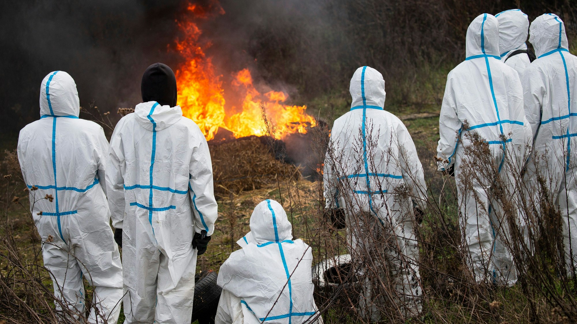 Aktivistinnen und Aktivisten und weißen Maler-Anzügen schauen auf einen brennenden Strohballen.