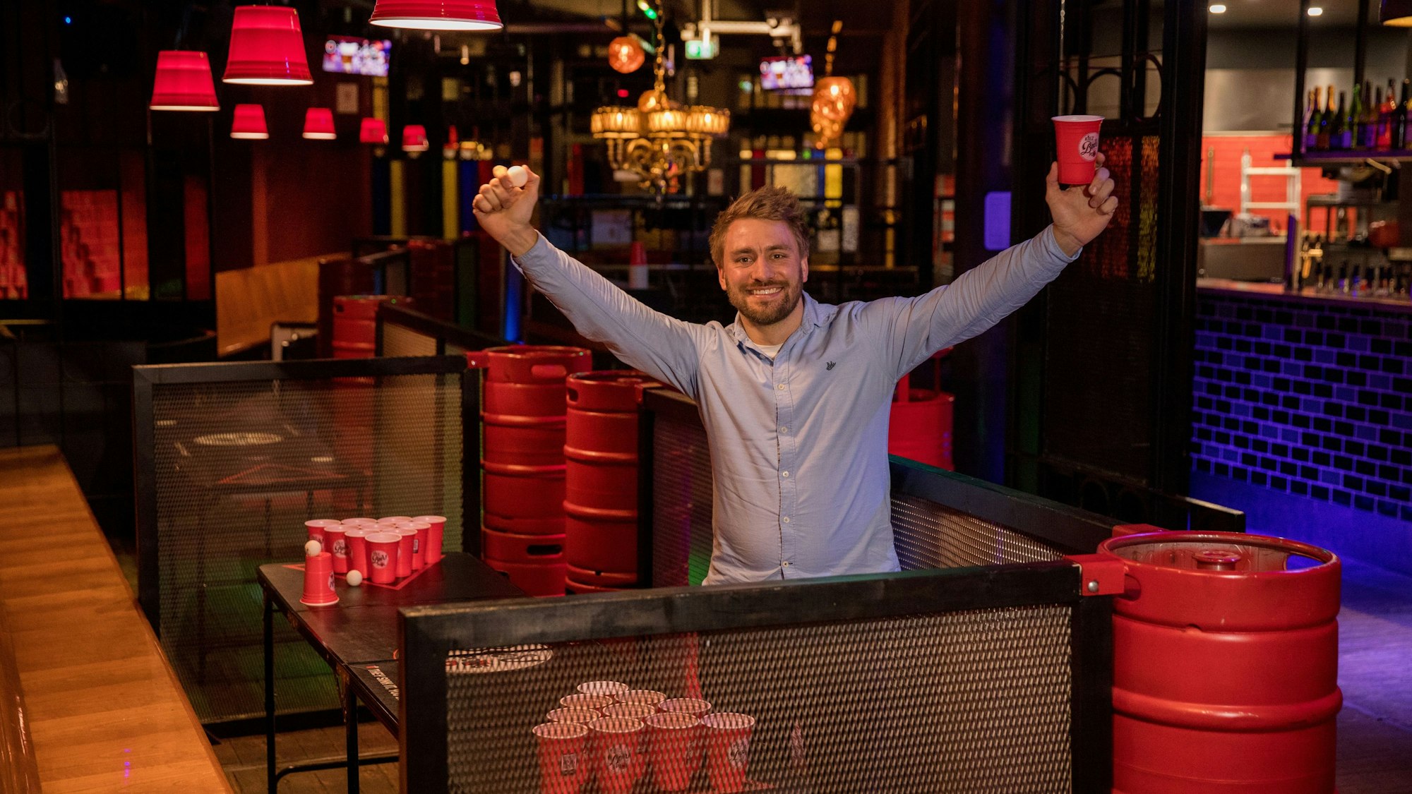 Marcel Bethke hält einen roten Beerpongbecher und einen Tischtennisball hoch. Er steht in seiner Bar vor einem Beerpongtisch.