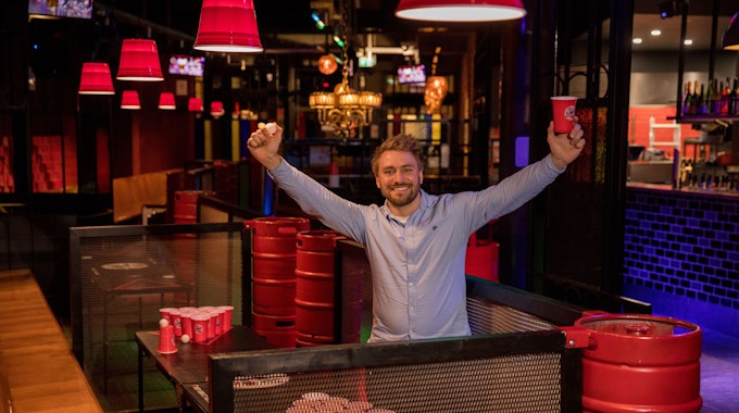 Marcel Bethke hält einen roten Beerpongbecher und einen Tischtennisball hoch. Er steht in seiner Bar vor einem Beerpongtisch.&nbsp;