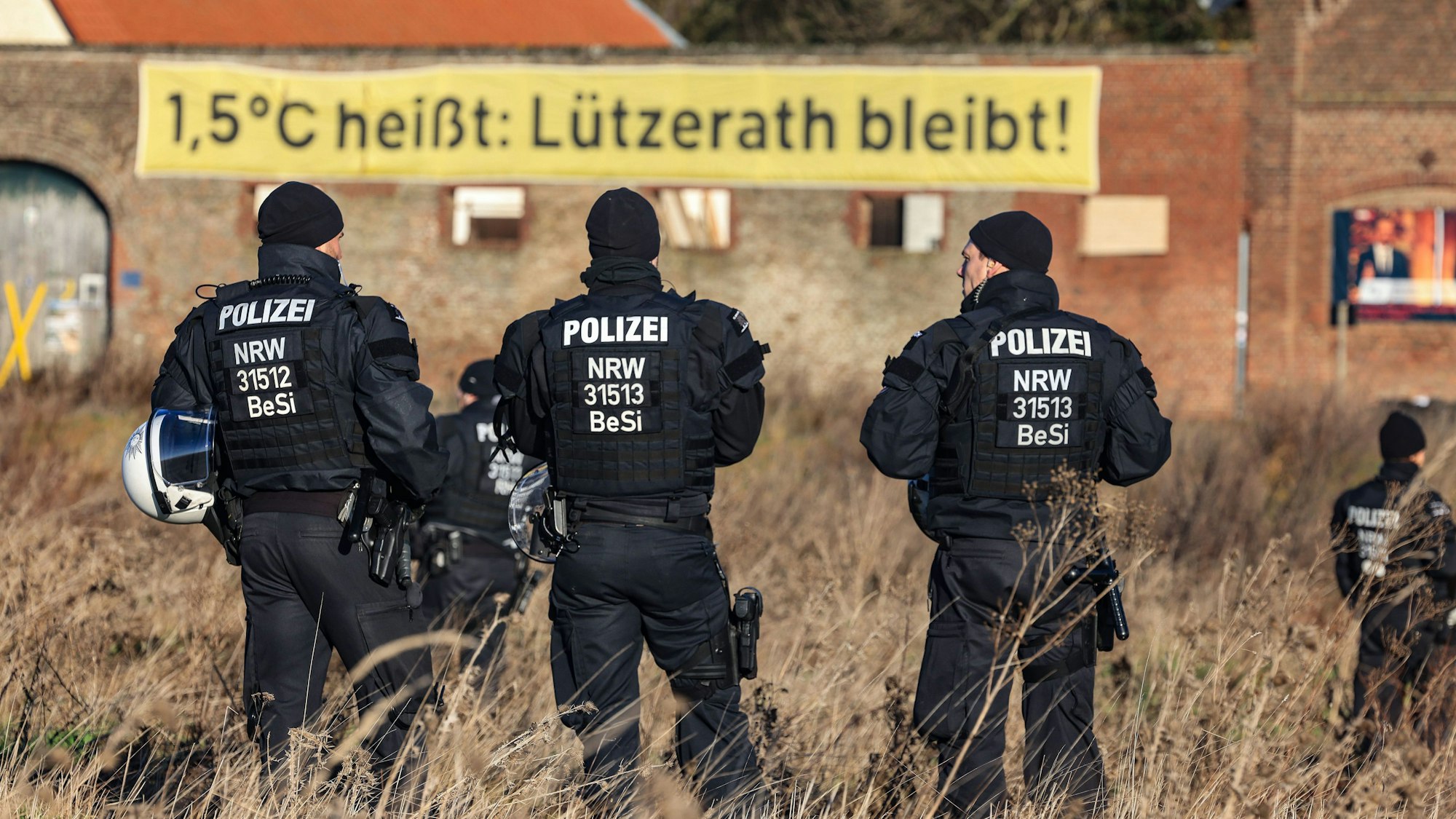 Polizisten stehen in Lützerath am Rande des Braunkohletagebaus, im Hintergrund hängt ein Banner „1,5°C heißt:Lützerath bleibt!“
