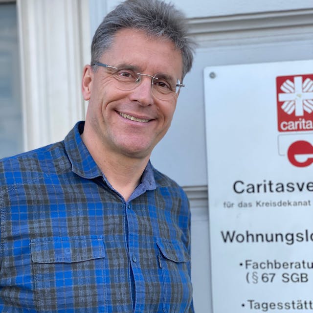Ralf Klaes steht vor einer Wand, an der ein Schild der Caritas zu erkennen ist.