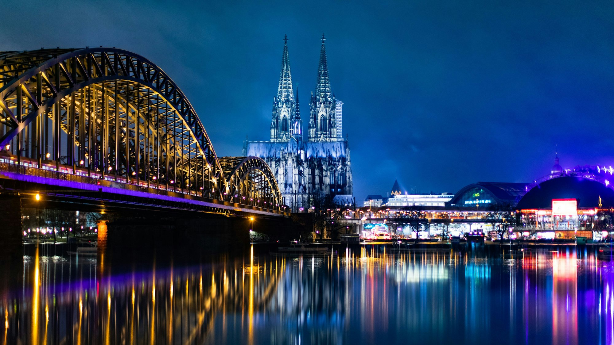 Hohenzollernbrücke, Kölner Dom (m) und der Musical Dome (r) spiegeln sich bei blauer Stunde in Regenwasser auf einer Mauer.