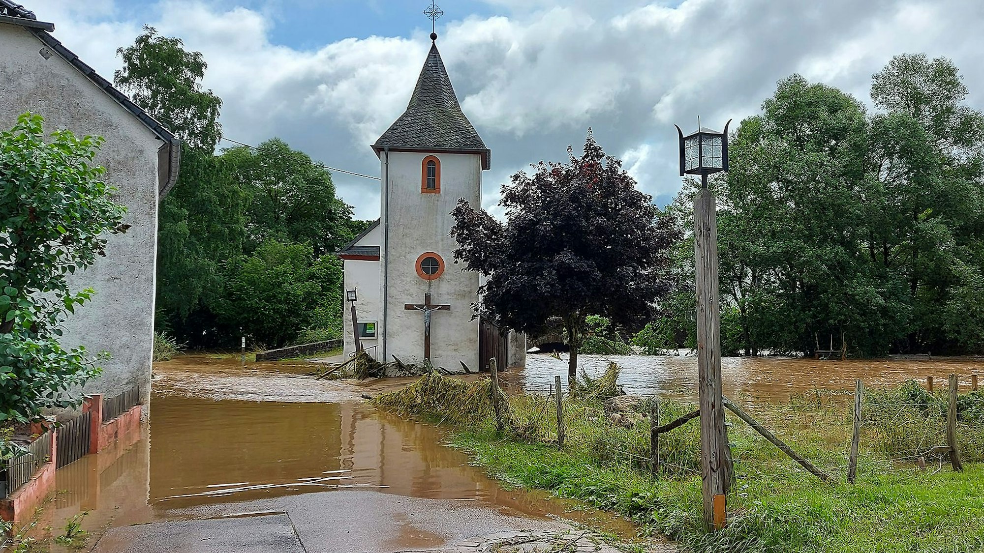 Hochwasser bedeckt eine Straße. Ein Haus und eine Kirche stehen im Wasser. Nur ein wenig Wiese kommt an der Seite des Bildes noch aus dem Wasser.