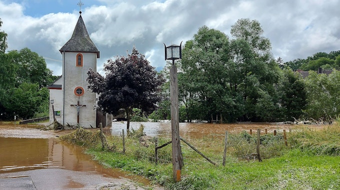 Hochwasser bedeckt eine Straße. Ein Haus und eine Kirche stehen im Wasser. Nur ein wenig Wiese kommt an der Seite des Bildes noch aus dem Wasser.