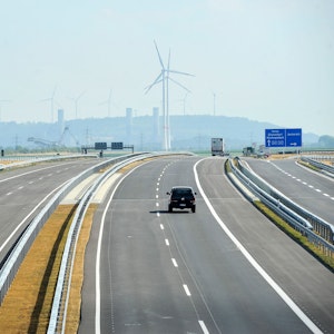 Ein Auto fährt auf einer Autobahn. Im Hintergrund sind Windräder und der obere Teil eines Braunkohle-Baggers zu sehen.