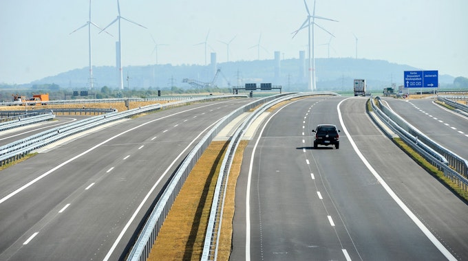 Ein Auto fährt auf einer Autobahn. Im Hintergrund sind Windräder und der obere Teil eines Braunkohle-Baggers zu sehen.
