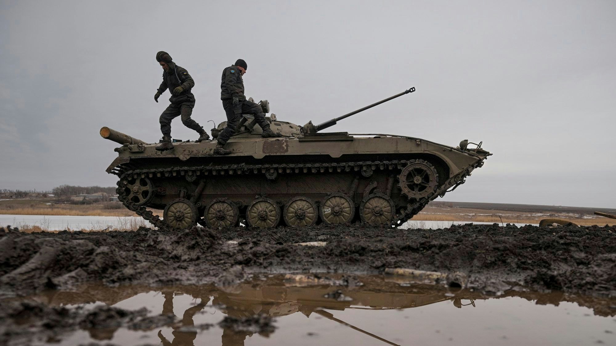 Zwei ukrainische Soldaten stehen während einer Schießübung auf einem Panzer, der auf einem Feld voller Pfützen steht.