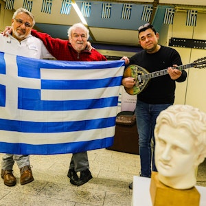 Kostas Giovanoglou (l.) und Vissarion Klissouras (M.) halten eine Griechische Flagge in der Hand, daneben steht Piros Dimitrios mit einem Musikinstrument namens Bouzouki.