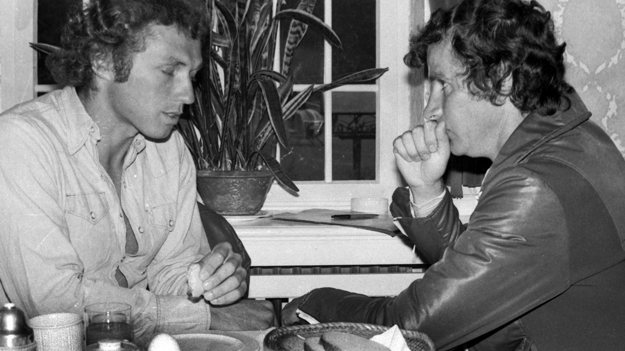 Zwei Männer sitzen sitz gegenüber und scheinen sich zu unterhalten. Im Vordergrund steht ein gedeckter Frühstückstisch mit gekochtem Ei und Brot. Das Foto ist in schwarz-weiß.