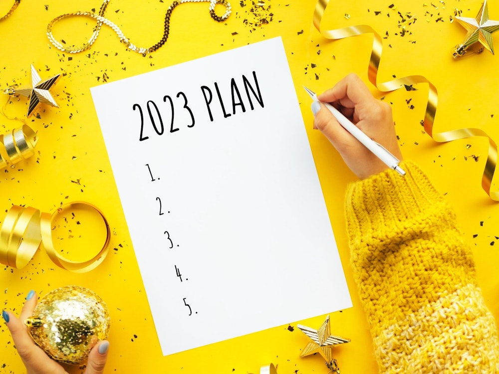 Eine Person schreibt einen Plan für 2023. Weihnachtsdekoration liegt um das Blatt Papier herum auf einem gelben Untergrund. Neben dem Papier ruht die rechte Hand der schreibenden Person mit einem weißen Stift in der Hand.