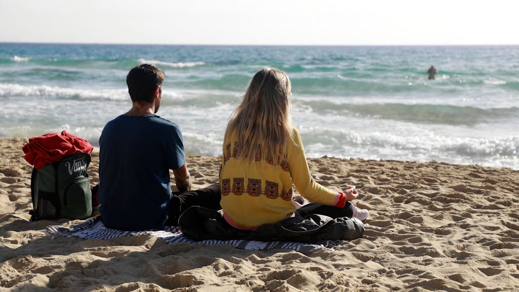 Das Foto zeigt zwei Menschen, die auf einer Decke am Strand sitzen und auf das Meer schauen.
