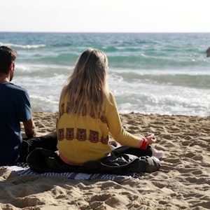 Das Foto zeigt zwei Menschen, die auf einer Decke am Strand sitzen und auf das Meer schauen.