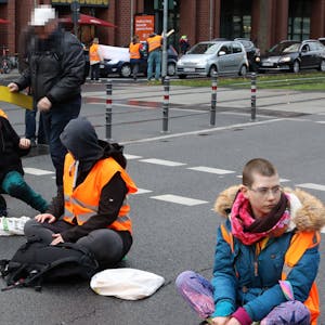 Aktivistinnen der „Letzten Generation“&nbsp;sitzen auf der Fahrbahn der Cäcilienstraße in Köln. Ein Passant versucht, die Aktion zu unterbinden.