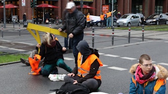Aktivistinnen der „Letzten Generation“sitzen auf der Fahrbahn der Cäcilienstraße in Köln. Ein Passant versucht, die Aktion zu unterbinden.