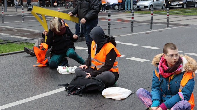 Aktivistinnen der „Letzten Generation“&nbsp;sitzen auf der Fahrbahn der Cäcilienstraße in Köln. Ein Passant versucht, die Aktion zu unterbinden.