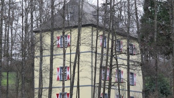 Die Burg Welterode in Eitorf mit gelber Fassade und weiß-roten Fensterläden, umringt von Bäumen.