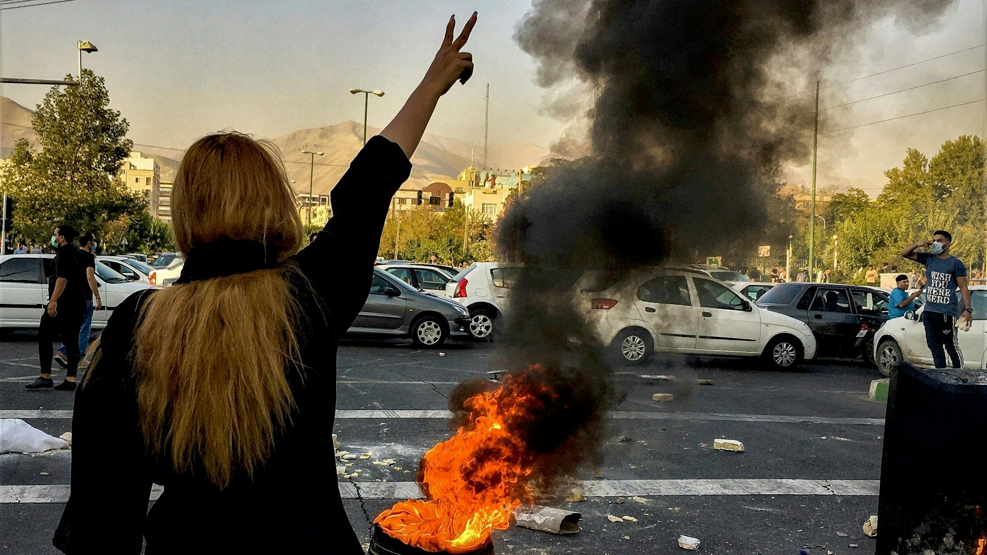 Eine Person mit langen Haaren steht vor einem brennenden Autoreifen und zeigt mit ihrer Hand das Victory-Zeichen.
