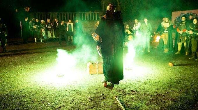 Ein Mann in Magierkostüm steht vor einer Kiste, neben ihm steigen grüne Feuerfontänen und Rauch empor.