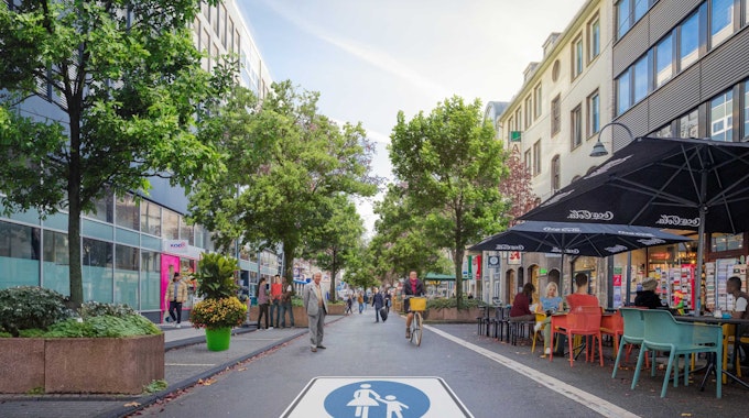 Auf der Straße prangt ein riesiges Piktogramm einer Fußgängerzone. Es sind Radfahrer und Fußgänger unterwegs, in einem Straßencafé sitzen Menschen.&nbsp;