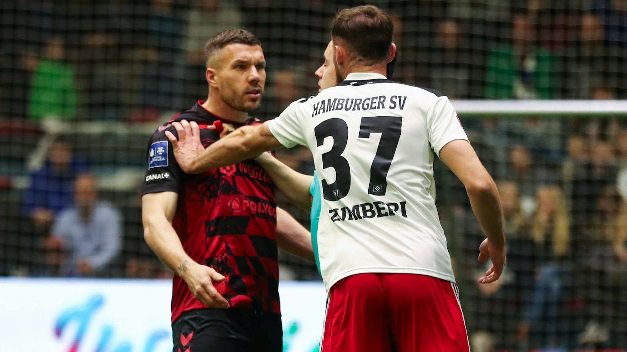 Lukas Podolski wird von einem Mann im weißen Trikot an der Schulter gefasst. Er schaut wütend.