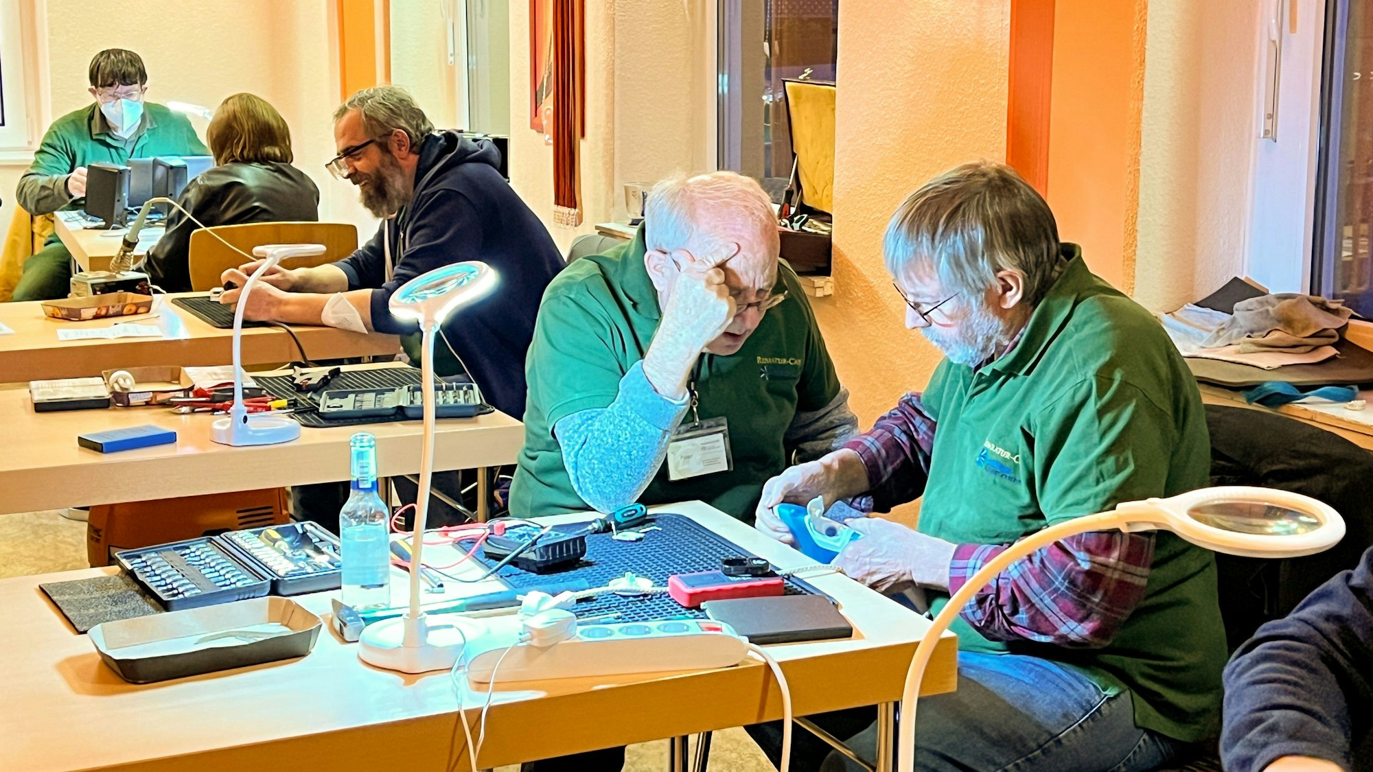 An mehreren Tischen sitzen ältere Männer und reparieren mit Werkzeug und unter Zuhilfenahme von Lupenlampen technische Geräte.