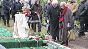 Eine Frau und ein Mann stehen vor einem offenen Grab und werfen Erde mit einer kleinen Schaufel hinein.