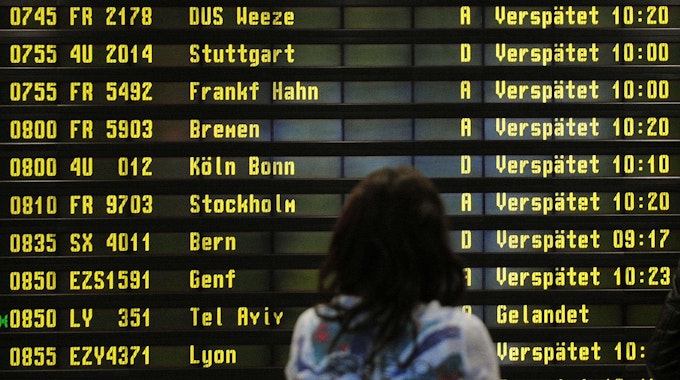 Eine Frau steht am Flughafen vor einer Anzeigetafel. Zahlreiche Flüge sind als "Verspätet" markiert.