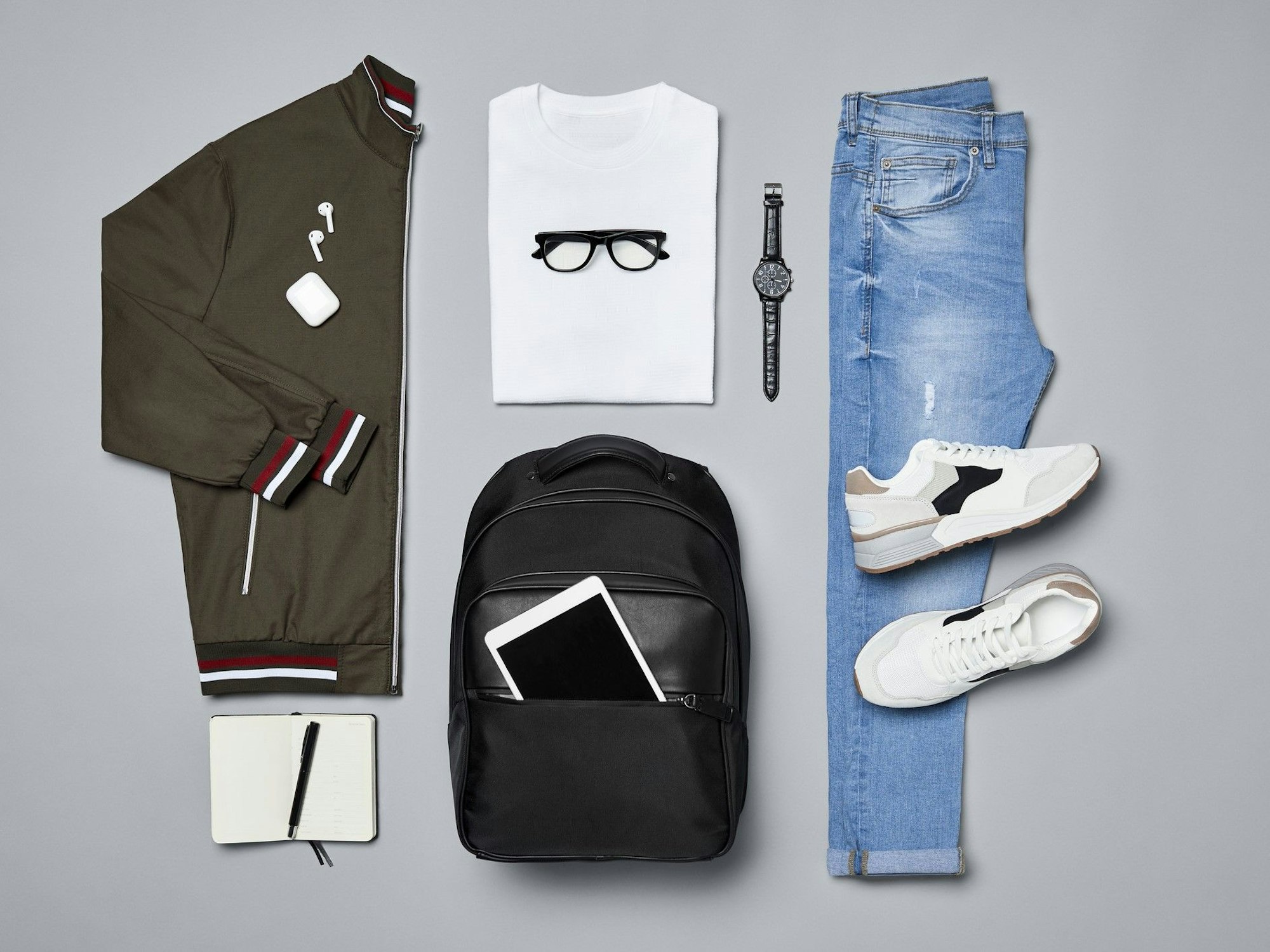 Verschiedene Kleidungsstücke und Accessoires wie eine Jacke, ein T-Shirt, eine Armbanduhr, ein Paar Schuhe, eine Jeans, ein Rucksack und ein iPad, ein Notizblock und ein Kugelschreiber liegen auf einem grauen Untergrund.