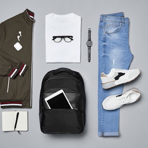Verschiedene Kleidungsstücke und Accessoires wie eine Jacke, ein T-Shirt, eine Armbanduhr, ein Paar Schuhe, eine Jeans, ein Rucksack und ein iPad, ein Notizblock und ein Kugelschreiber liegen auf einem grauen Untergrund.