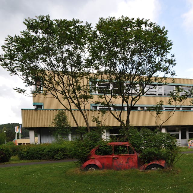 Das Schulzentrum Cyriax in Overath vor Bäumen, davor ein mit Pflanzen bewachsener roter Wagen.