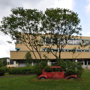 Das Schulzentrum Cyriax in Overath vor Bäumen, davor ein mit Pflanzen bewachsener roter Wagen.