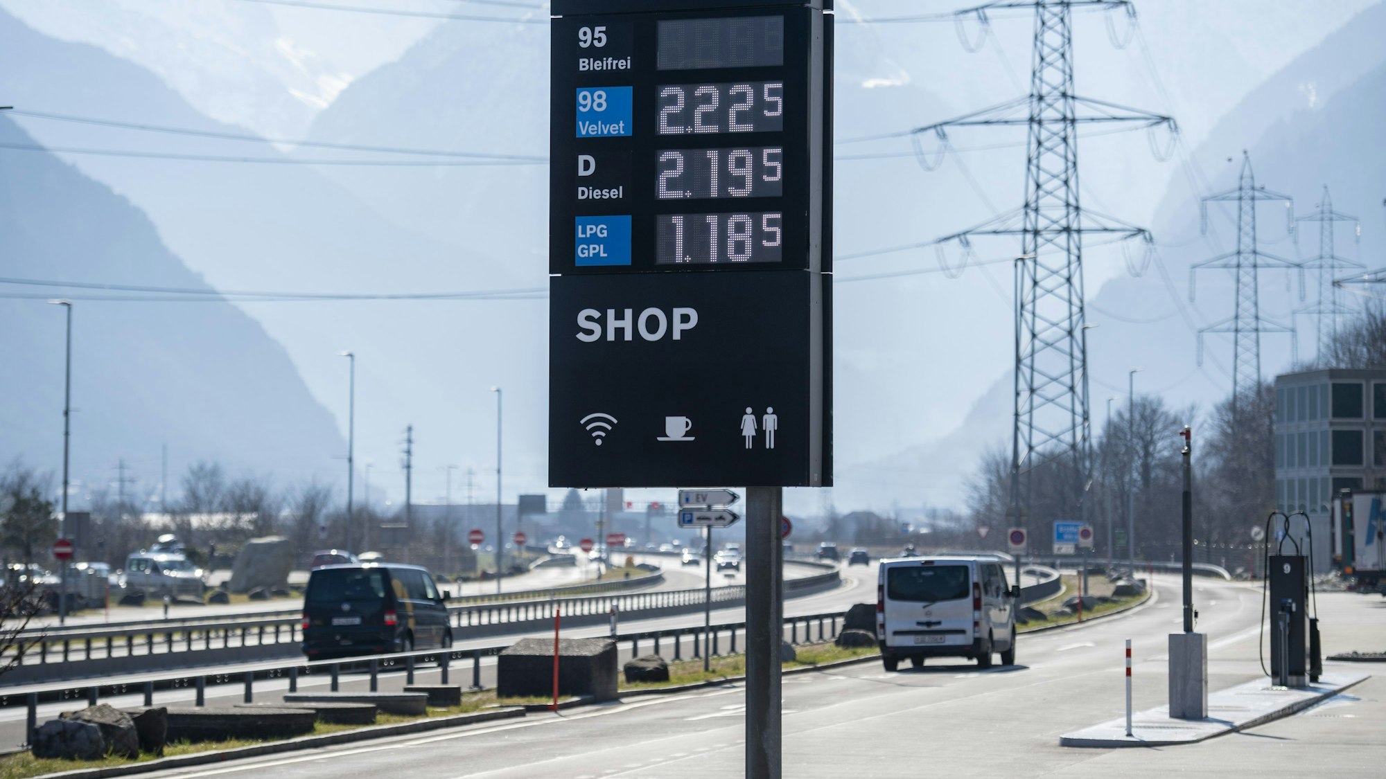 Eine Tankstelle an einer Autobahn. Im Vordergrund steht eine Preistafel für Kraftstoff, im Hintergrund fahren Autos.
