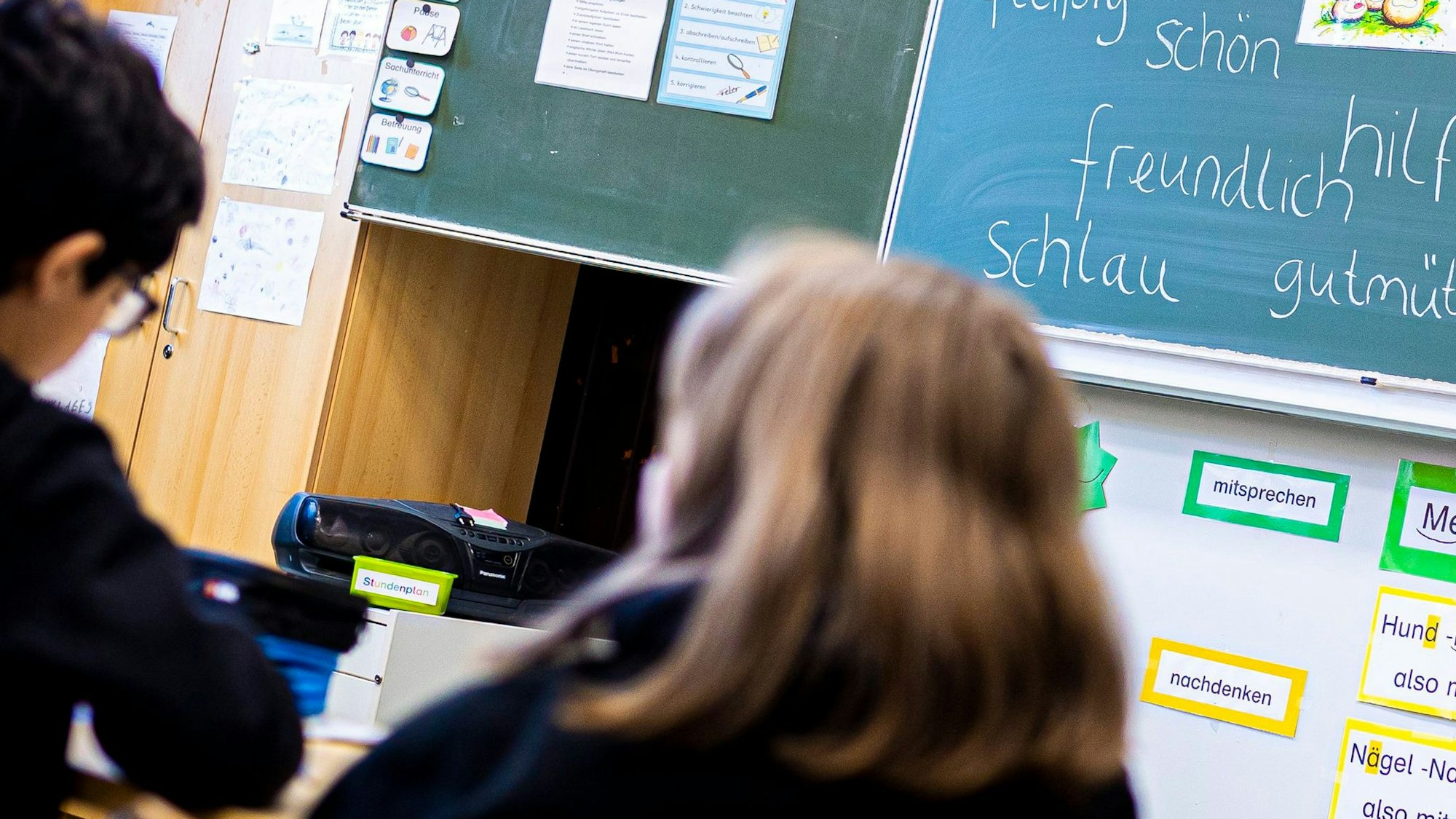 Drei Kinder in Schwarz sitzen in einem Klassenraum vor einer Tafel, die mit Adjektiven beschrieben ist.
