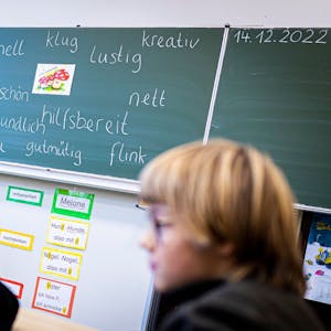 Drei Kinder in Schwarz sitzen in einem Klassenraum vor einer Tafel, die mit Adjektiven beschrieben ist.&nbsp;