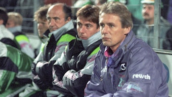 Jürgen Gelsdorf sitzt bei einem Spiel von Borussia Mönchengladbach im DFB-Pokal auf der Trainerbank und trägt einen Trainingsanzug. Neben ihm sitzt Bernd Krauss, sein damaliger Co-Trainer, in Trainingsjacke und mit verschränkten Armen.