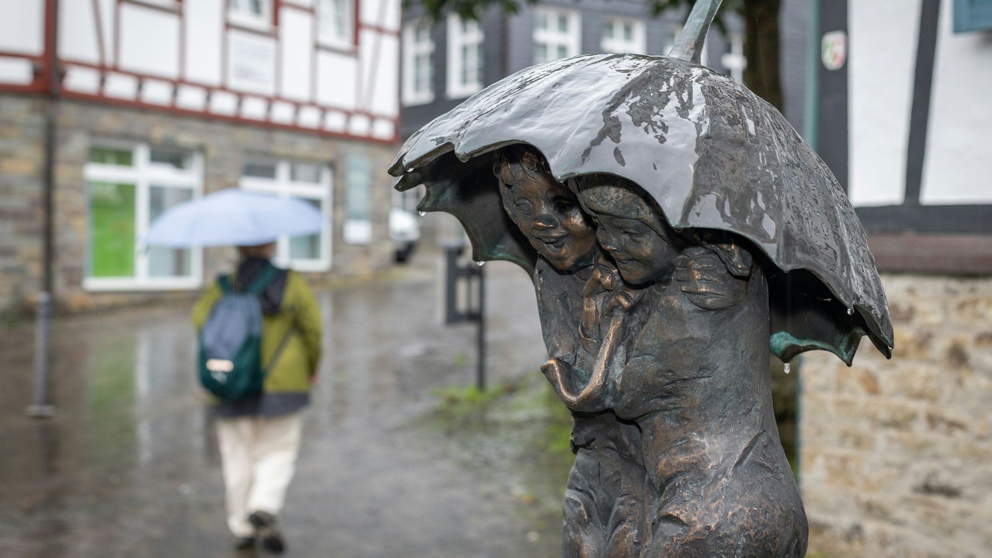 Eine Bronze-Statue aus zwei Personen hat einen Regenschirm über den Köpfen. Im Hintergrund geht eine Person mit Regenschirm.