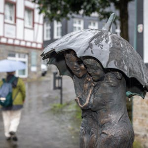 Eine Bronze-Statue aus zwei Personen hat einen Regenschirm über den Köpfen. Im Hintergrund geht eine Person mit Regenschirm.&nbsp;