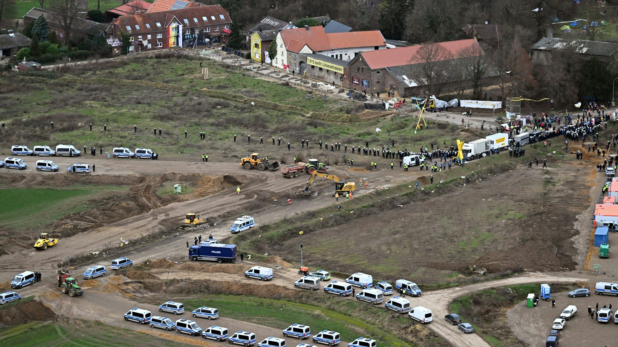 Polizeiautos und Polizisten stehen vor der Ortschaft Lützerath - eine Luftaufnahme zeigt die Szenerie. Lützerath soll zur Erweiterung des Braunkohletagebaus Garzweiler II abgebaggert werden.