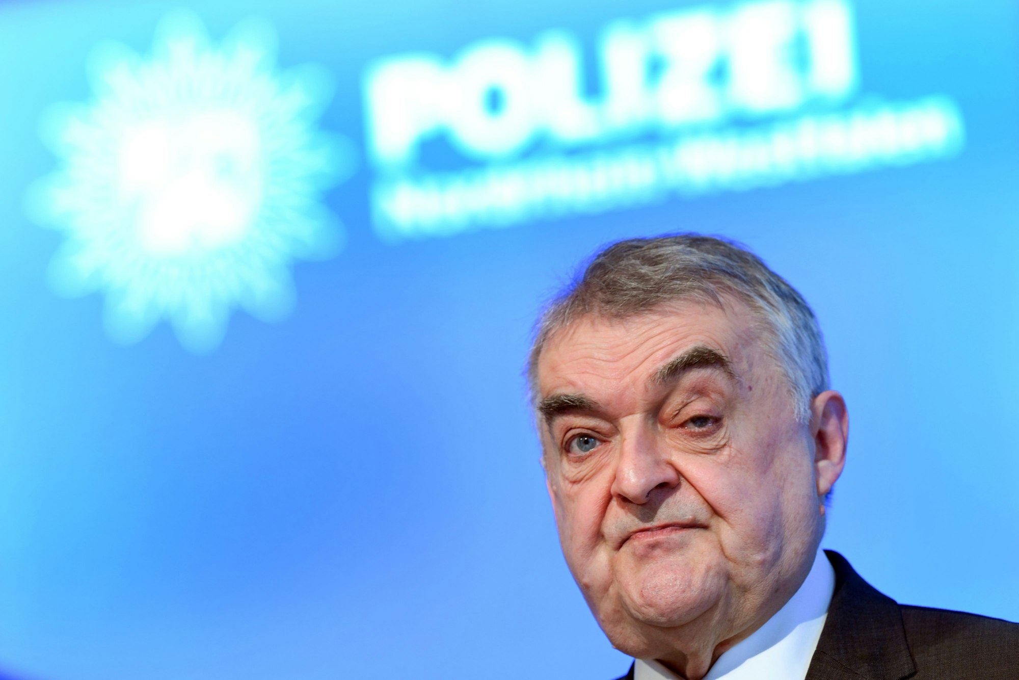 Innenminister Herbert Reul steht vor einer blauen Wand, auf der groß, unscharf im Hintergrund, das Wort „Polizei“ steht.