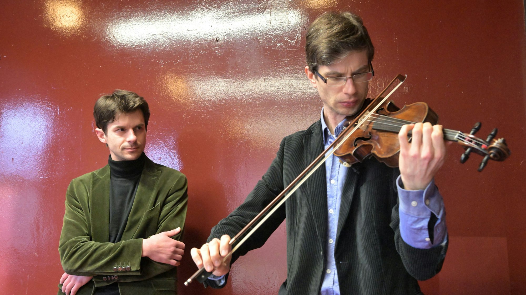Roman Salyutov steht an einer Wand hinter Alexander Lifland, der auf der historischen Geige spielt.