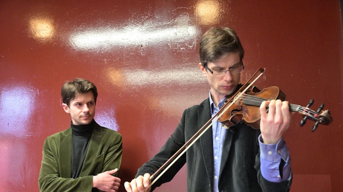 Roman Salyutov steht an einer Wand hinter Alexander Lifland, der auf der historischen Geige spielt.